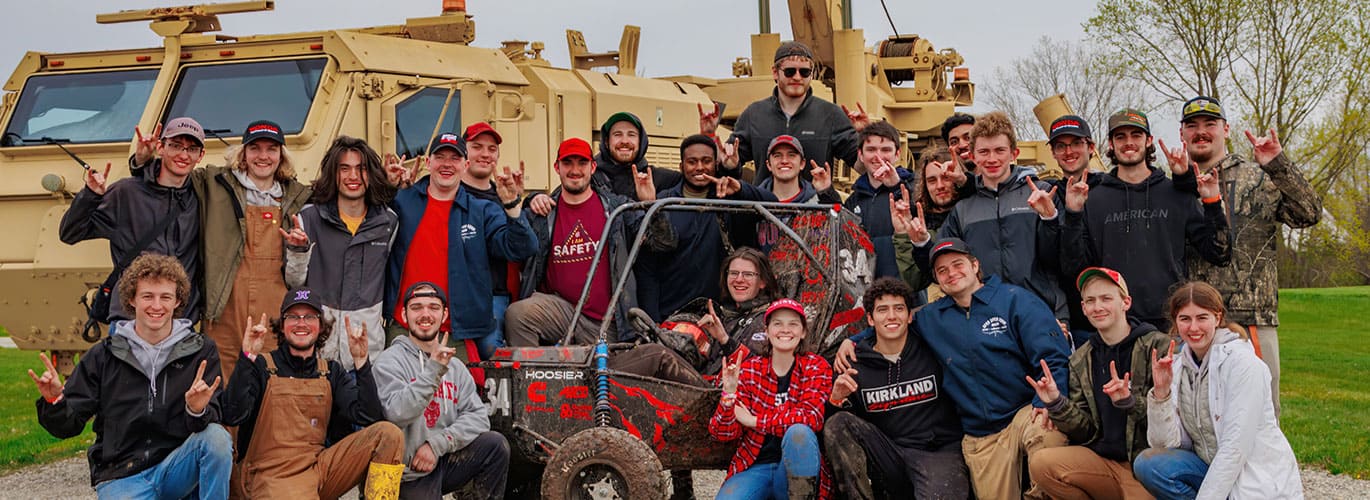 Group of joyful individuals posing in front of a military vehicle, with a muddy buggy in the foreground.