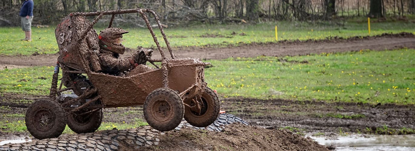 Mud-covered buggy in action on a rough terrain, with a spectator watching.