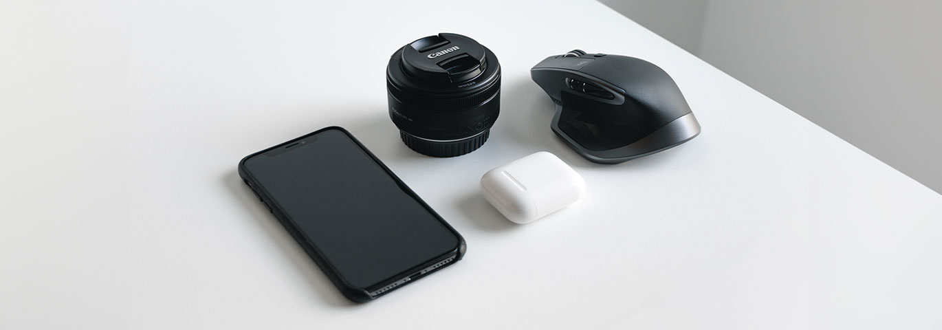 Smartphone, camera lens, earbuds case, and computer mouse on a white surface.