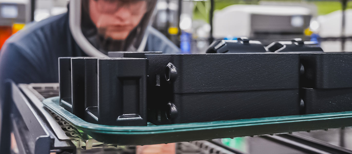 Close-up of black machinery part on green tray; blurry worker in the background.